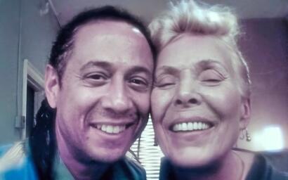 Two smiling individuals close together, one with dark hair and the other with short platinum blonde hair.