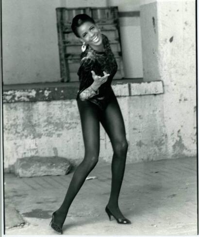A woman smiling and pointing at the camera, wearing a patterned long-sleeve top, tights, and high heels in an urban setting.