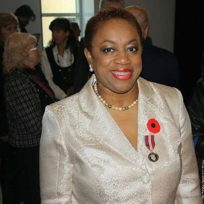 Elegant woman in a cream-colored suit with medals and a pearl necklace at a formal event.