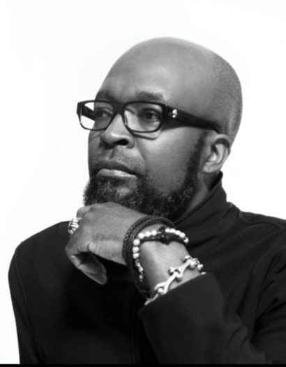 Black and white portrait of a man with glasses, a beard, and layered bracelets, posed thoughtfully.