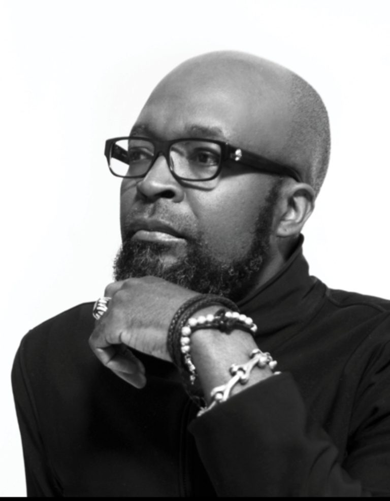 Black and white portrait of a man with glasses, a beard, and layered bracelets, posed thoughtfully.
