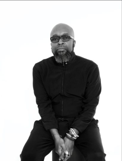 Black-and-white portrait of a man wearing glasses, a zip-up jacket, and bracelets.