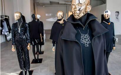 Mannequins dressed in black fashion outfits with gold mirrored masks in a gallery setting.