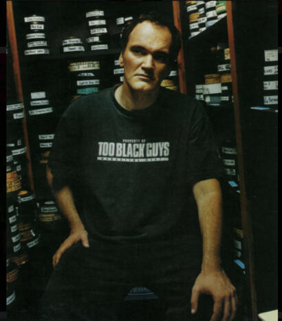 Man in a black T-shirt reading "Property of Too Black Guys," standing in front of shelves filled with labeled boxes.
