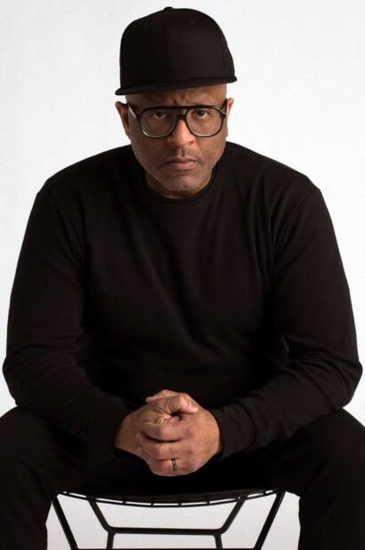 Man in black sweatshirt, cap, and glasses seated against a white background.