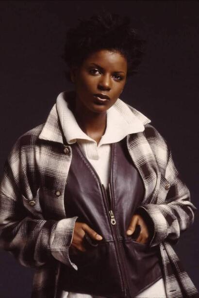 A woman wearing a plaid jacket over a leather vest and a white hoodie, styled with bold makeup.