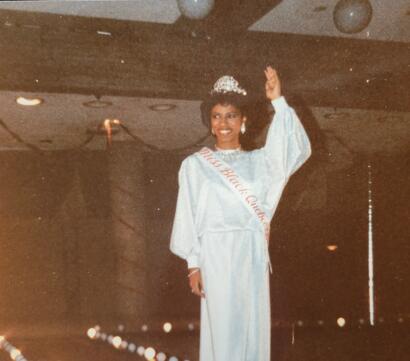 Miss Black Queen wearing a sash and crown, smiling and waving.