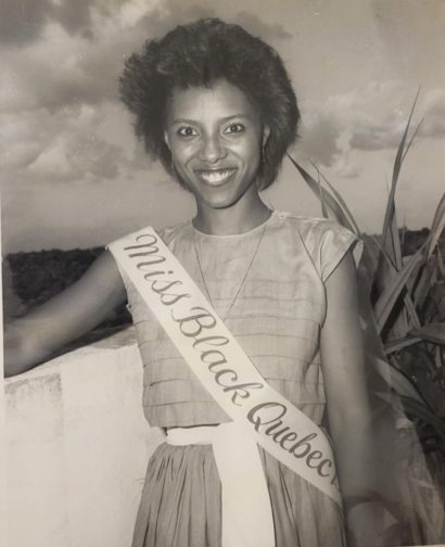 Young woman wearing a sash that reads “Miss Black Queen” smiles outdoors in a black and white photo.