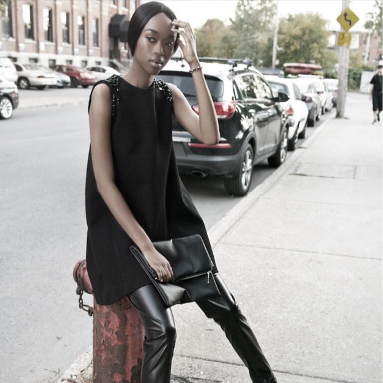 Fashion-forward woman in a black sleeveless top with embellished shoulders, leather pants, posing on city street.