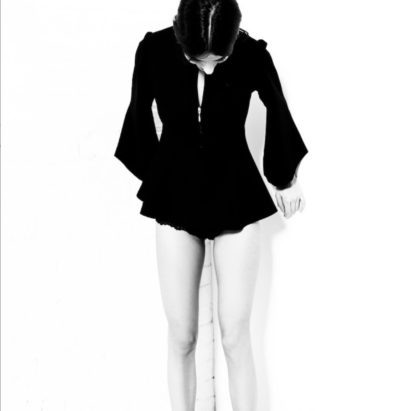 A black and white photo of a woman wearing a black top with bell sleeves, looking downward, and standing against a white background.