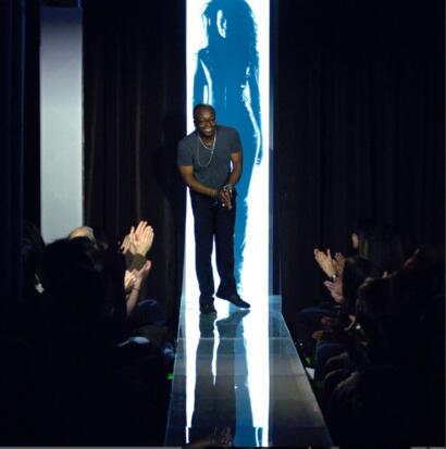 A fashion show runway with a designer greeting the audience, framed by a blue-lit poster of a model.