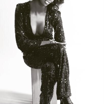 Woman in a sparkling, sequined jumpsuit sitting on a wooden stool, smiling with confidence.