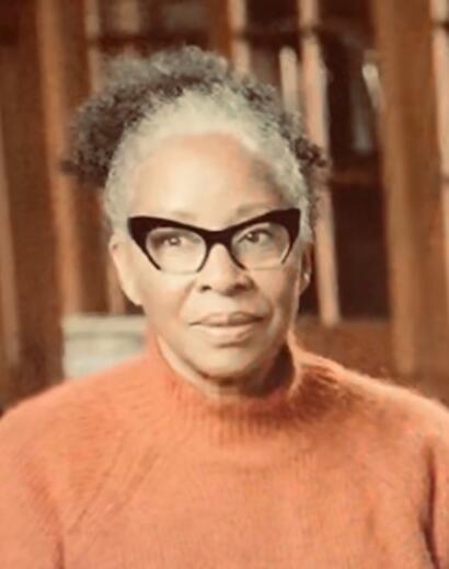 Portrait of a woman wearing black glasses and an orange sweater, indoors with bookshelves in the background.