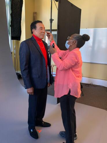 A man in a blazer receives makeup application from a stylist in a studio setting.