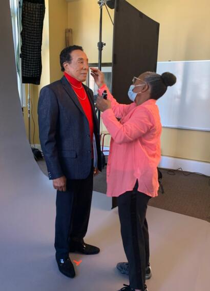 A man in a blazer receives makeup application from a stylist in a studio setting.