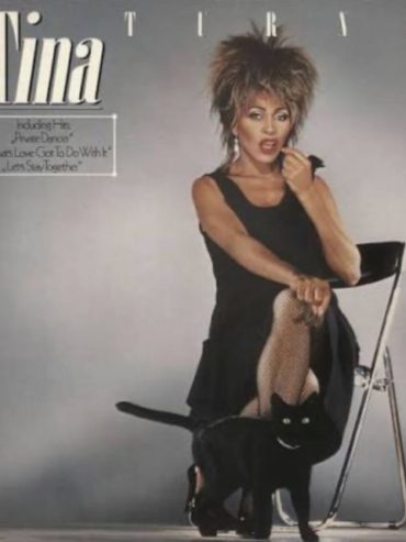 Tina Turner poses in a stylish black outfit with her signature energetic vibe, accompanied by a black cat.