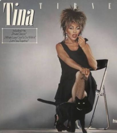 Tina Turner poses in a stylish black outfit with her signature energetic vibe, accompanied by a black cat.