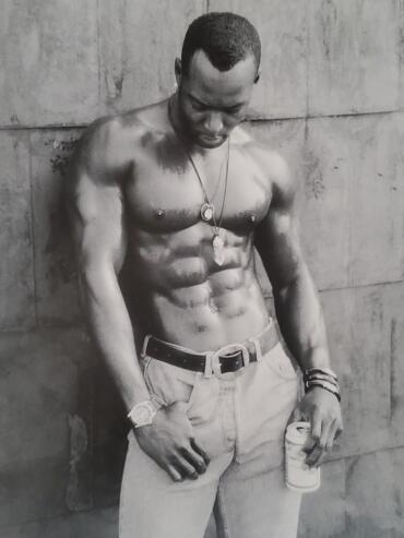 Black-and-white photo of a shirtless man with a muscular physique, wearing jeans, watch, and jewelry.
