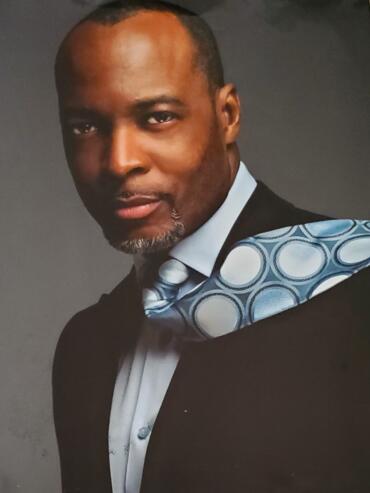 Formal portrait of a man in a black suit, white shirt, and patterned tie, against a dark background.
