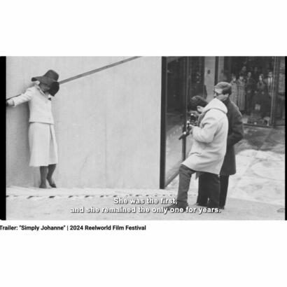 Black-and-white photo of two men filming a woman in a vintage coat and wide-brimmed hat outside a building.