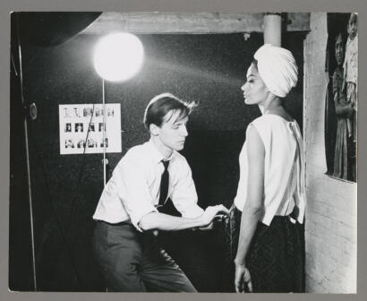 Black-and-white photo of a fashion model with a headwrap posing as a stylist adjusts her outfit, with a stylist working on her appearance.