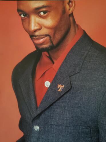 Close-up portrait of a man in a tailored dark blazer with lapel pin and red shirt, against a warm, reddish background.