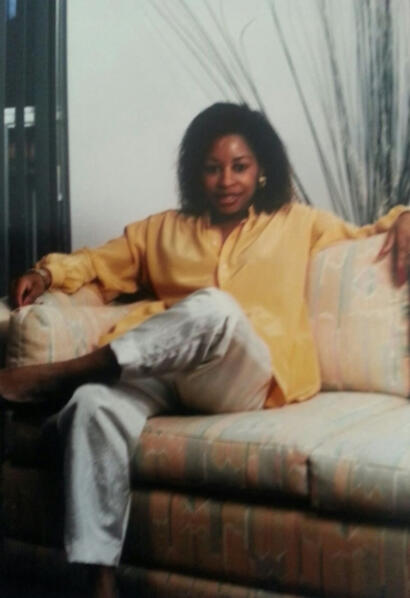 Woman with curly hair in a yellow blouse, seated on a patterned sofa with a relaxed pose.