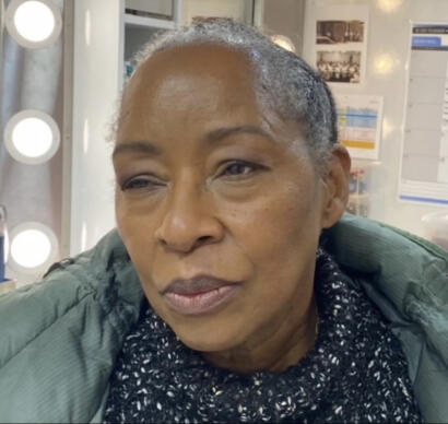 Close-up of a woman with short, graying hair, wearing a dark patterned scarf and green jacket, in a bright indoor setting.