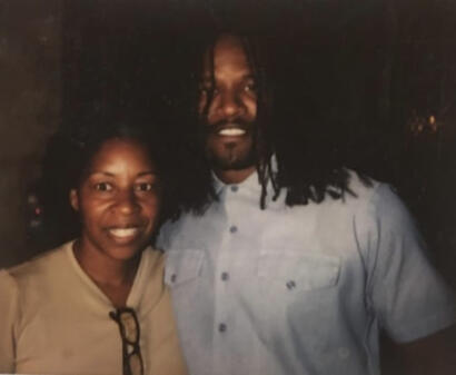 Two individuals pose together, one with glasses and a beige shirt, the other with long dreadlocks and a light blue shirt.
