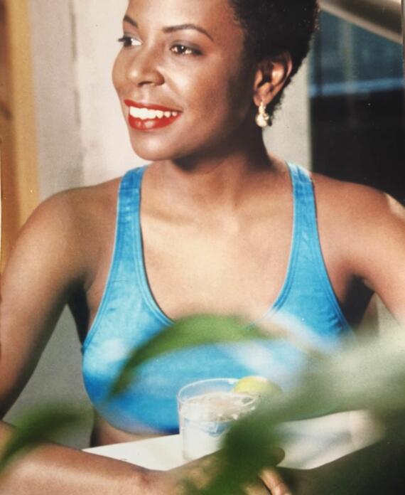 Woman smiling in a blue tank top with red lipstick, jewelry, and short hair.