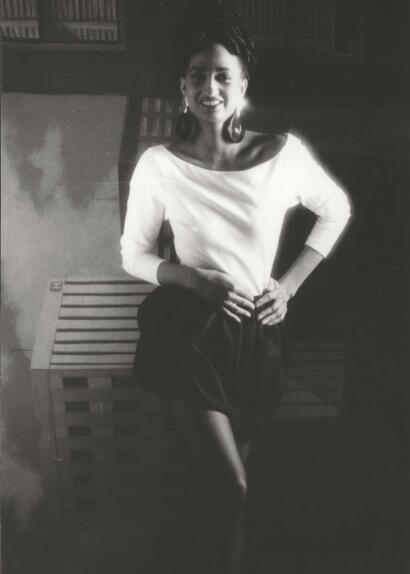 A woman in stylish attire, seated confidently against a bookshelf background.