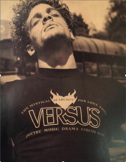 A young man with curly hair tilts his head back, wearing a black shirt with "Versus" branding and promotional text.