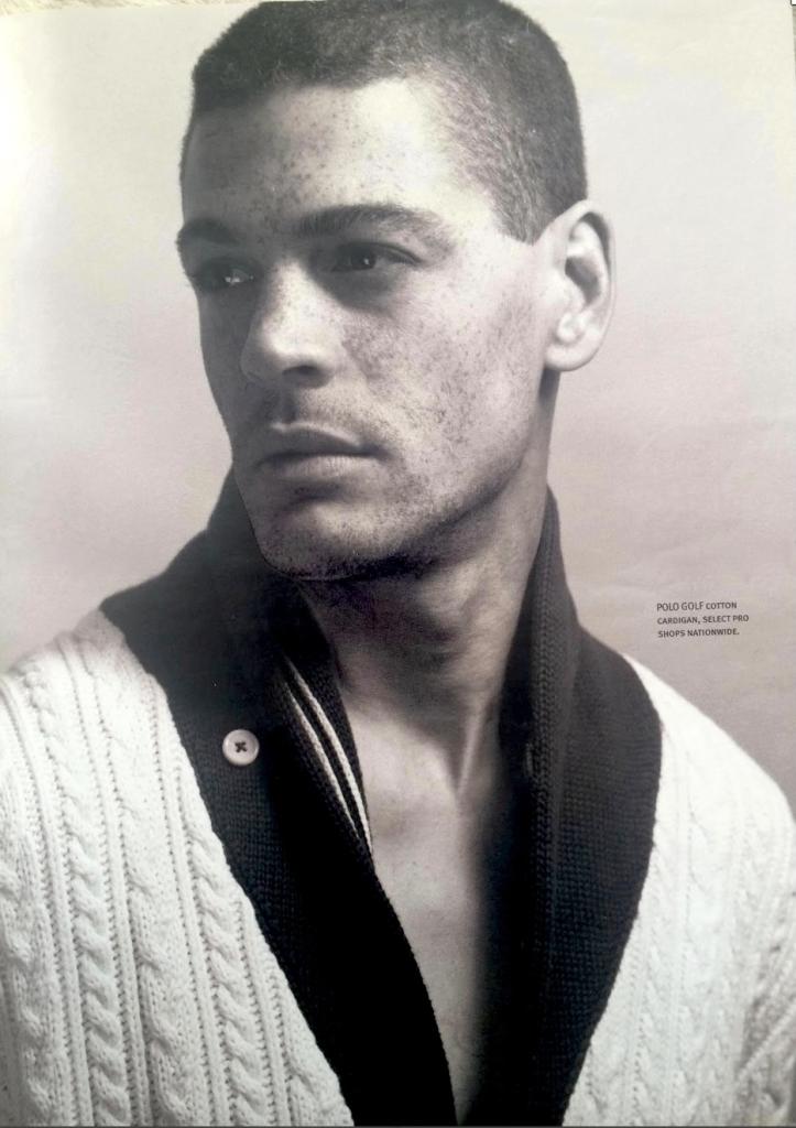 Black-and-white fashion portrait of a young male model wearing a cable-knit sweater and varsity jacket.