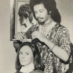 Black-and-white photo of a barber styling a woman's hair, with a fashion poster in the background.