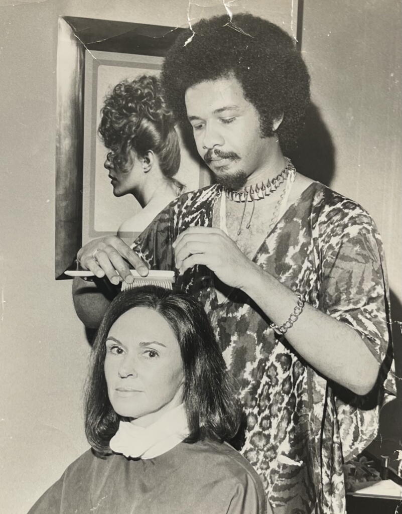 Black-and-white photo of a barber styling a woman's hair, with a fashion poster in the background.