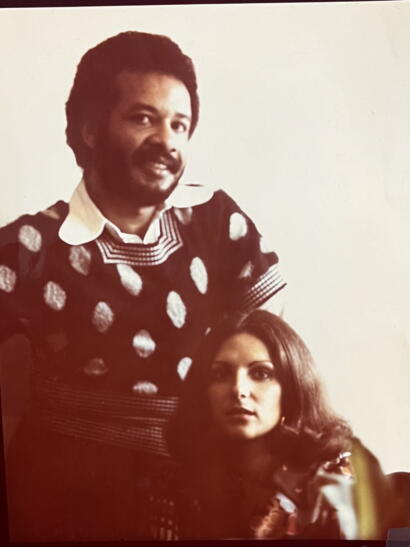 A man with a mustache and curly hair, dressed in a patterned sweater, seated beside a woman with voluminous hair and a thoughtful expression.