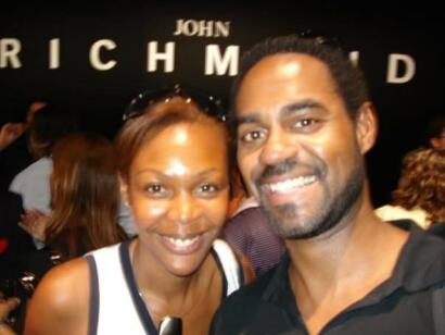 Two smiling individuals taking a selfie at a fashion event with "John Richard" branding in the background.