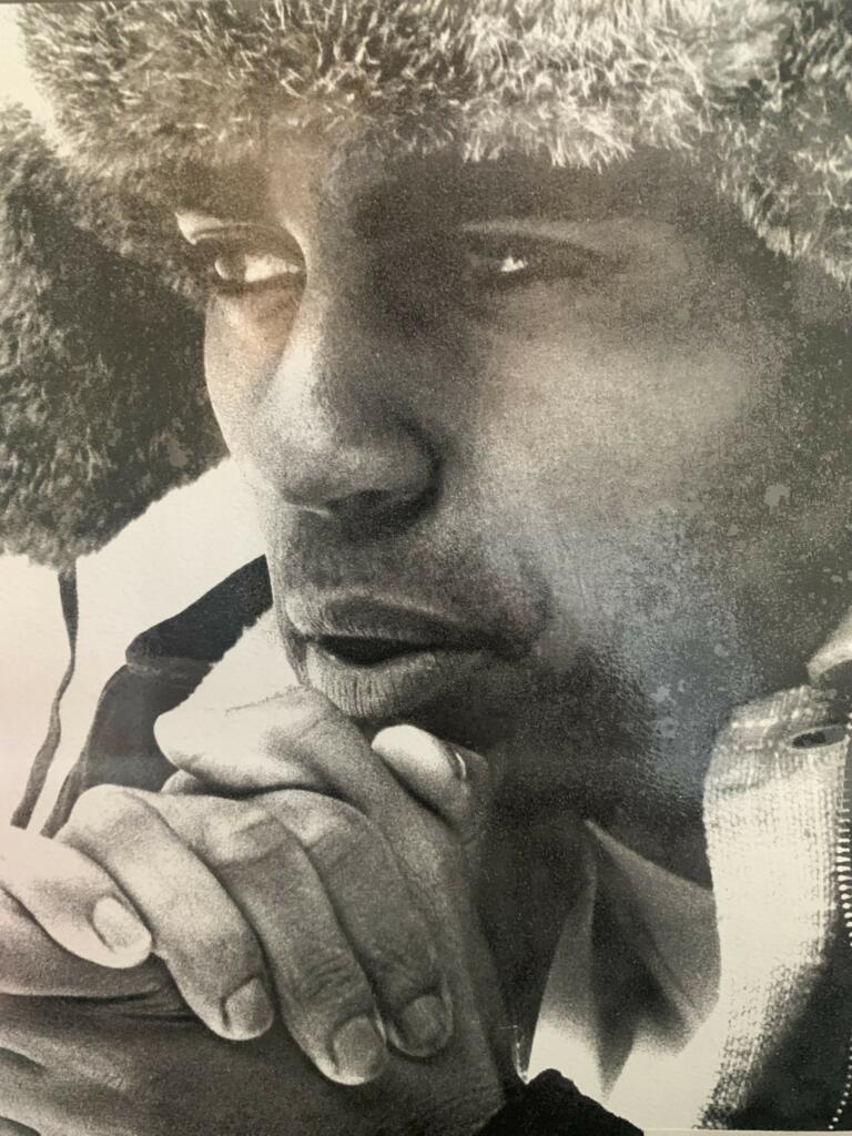 Close-up portrait of a person with textured hair, resting their chin on intertwined hands.