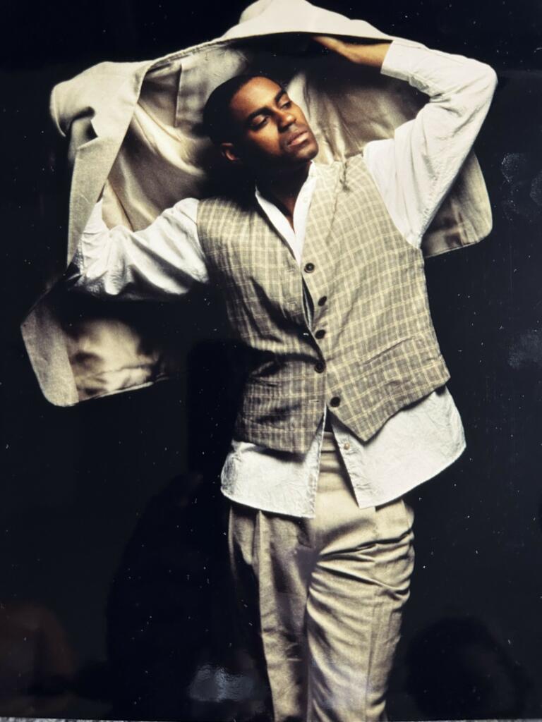 Man with closed eyes, dressed in layered neutral-toned clothing, posing against a dark background.