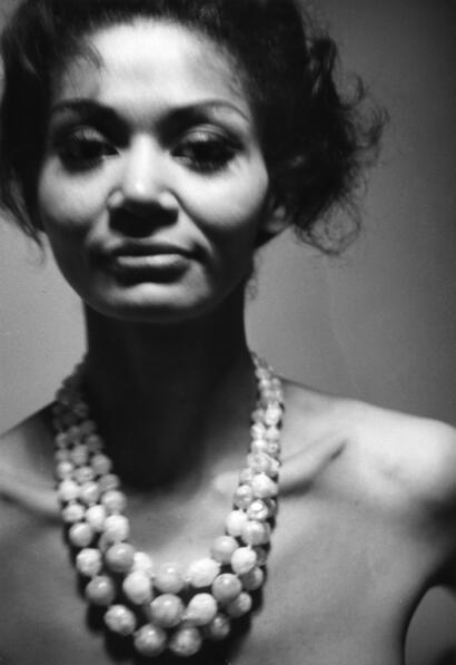 Black and white portrait of a woman with short, curly hair wearing a layered pearl necklace.