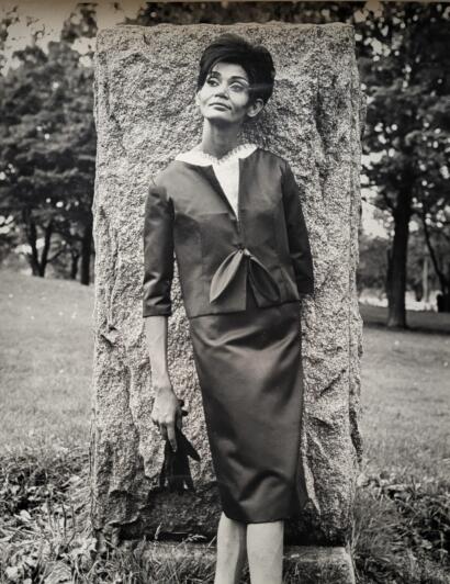 Black-and-white photo of a stylish woman in vintage fashion, standing outdoors next to a large stone.