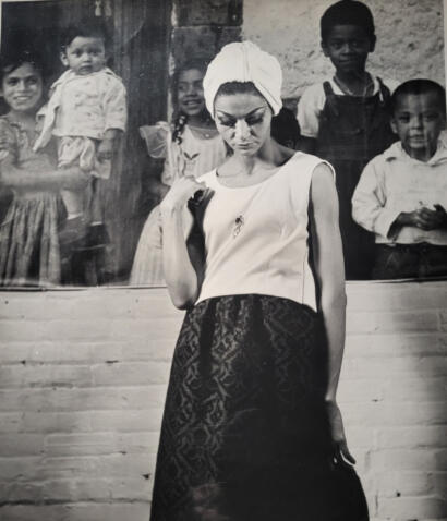 A woman with a headwrap and stylish outfit stands in front of a colorful mural of children.