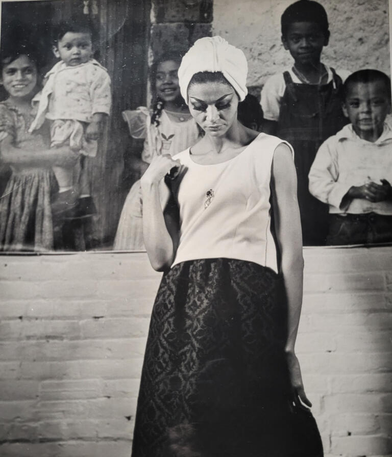 A woman with a headwrap and stylish outfit stands in front of a colorful mural of children.