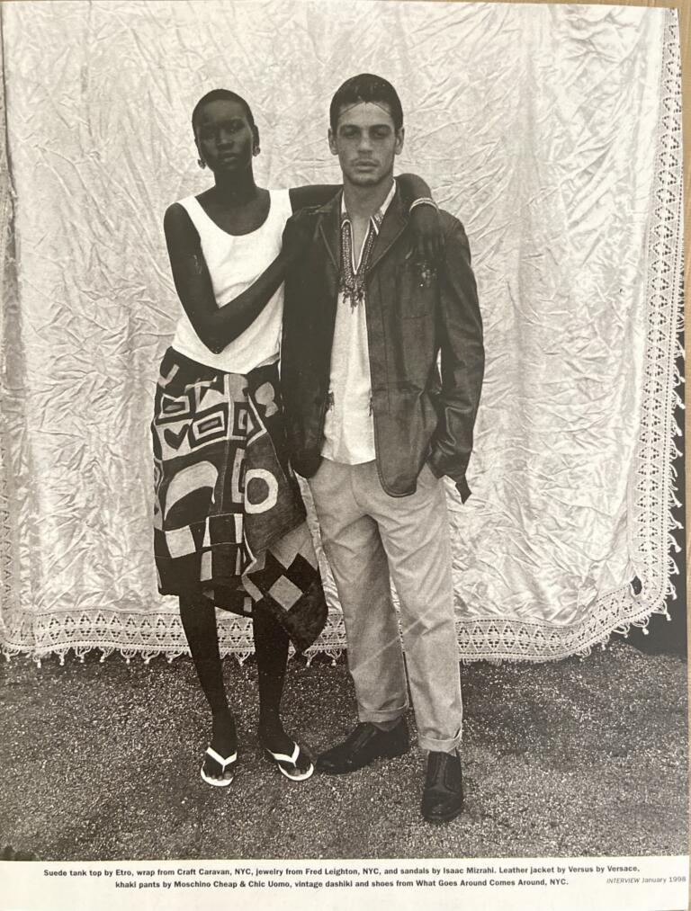 A stylish duo posed against a textured backdrop, showcasing 1998 fashion with jewelry, leather, and bold patterns.