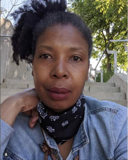 Close-up portrait of a woman with natural curly hair, wearing a denim jacket and bandana, outdoors with trees in the background.