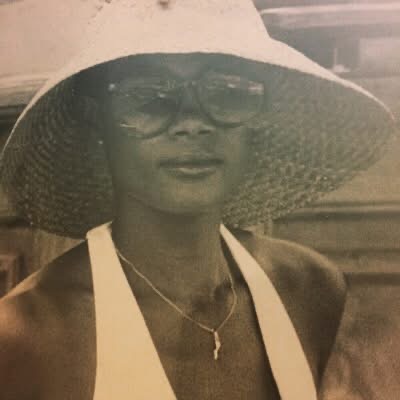 Woman in large sun hat, sunglasses, and white top, posing outdoors.