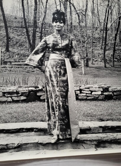 Elegant woman in a patterned, long dress with a sash, standing outdoors among trees.