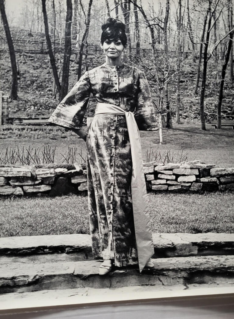 Elegant woman in a patterned, long dress with a sash, standing outdoors among trees.