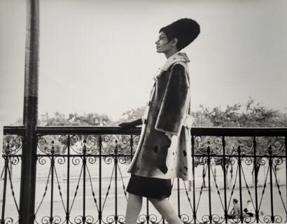 Black-and-white image of a fashionable woman standing by a decorative iron railing outdoors.
