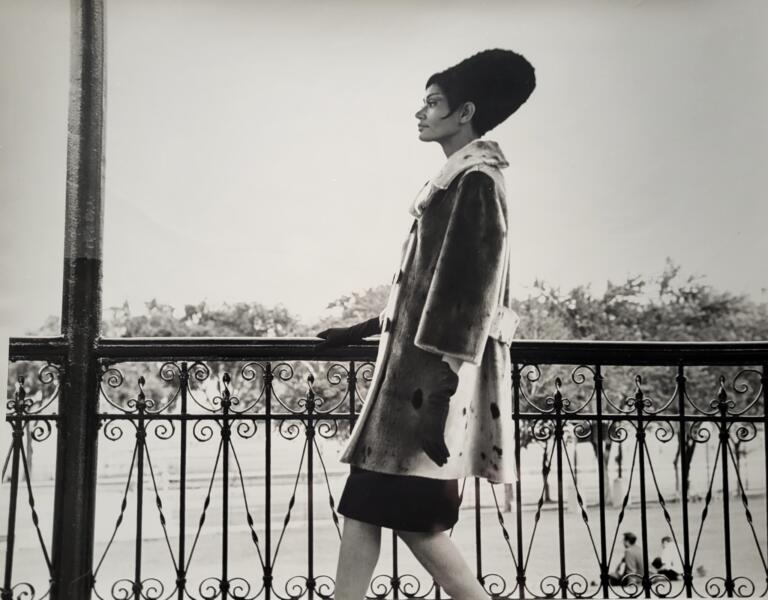 Black-and-white image of a fashionable woman standing by a decorative iron railing outdoors.